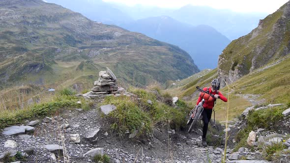 A man walking with his mountain bike on a trail in a European mountain range alt