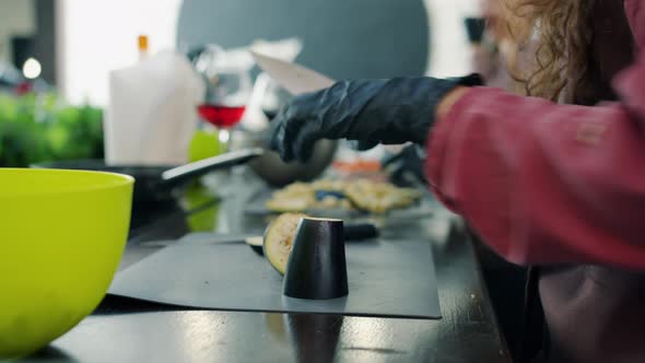 Close-up of Female Hands in Gloves Cutting Eggplant in Kitchen Cooking Food alt