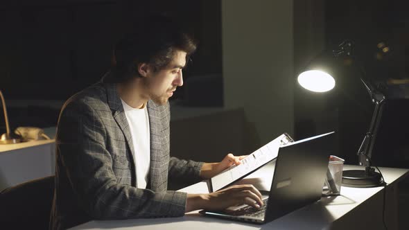 A Tired Man is Looking at a Financial Report on a Computer in the Office at Night