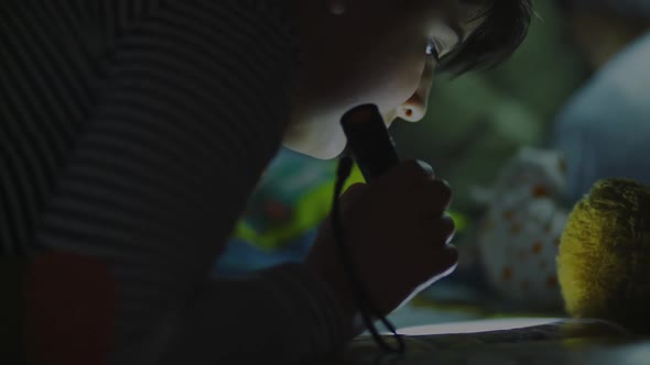 Boy Reads with Flashlight Under Blanket with Toys