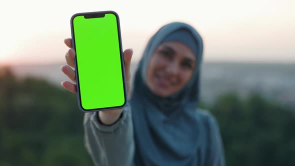 Beautiful Woman in Hijab on Top of the Hill in the Evening Keeps Smartphone with Green Screen on It
