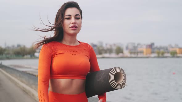 Beautiful woman goes after workout outdoors. The girl carries a fitness mat in her hands and smiles.