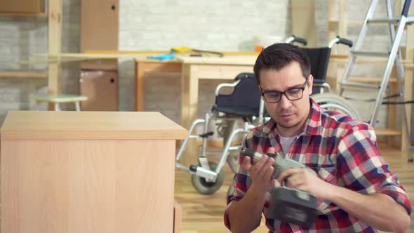 Portrait of a Young Disabled Man Next To a Wheelchair Collects a Bedside Table alt