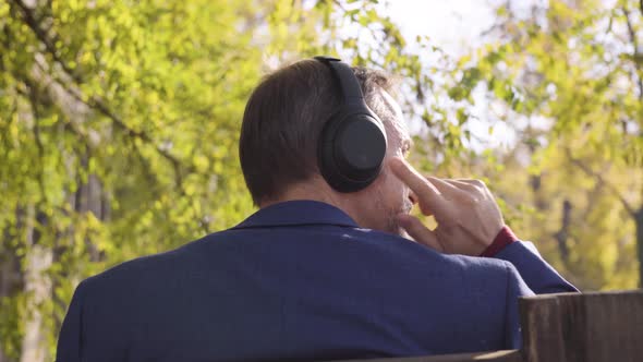 A Middleaged Handsome Caucasian Man Listens to Music with Headphones As He Sits on a Bench