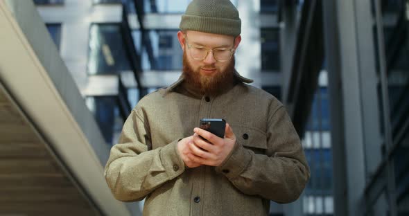A Bearded Man is Typing on a Mobile Phone While Standing Near Office Building alt