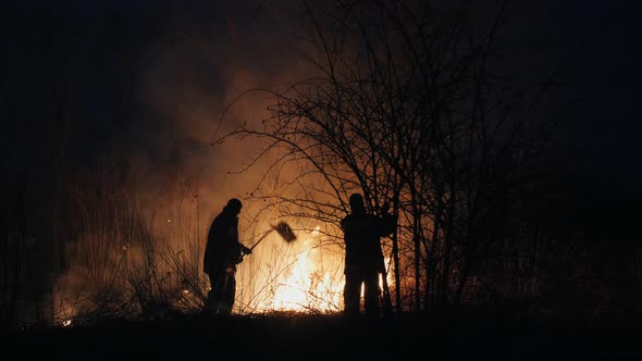 Two Firefighters with Fire Flappers Extinguish a Fire in the Forest at Night alt