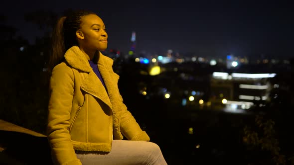 A Young Black Woman Waves at the Camera with a Smile As She Sits in an Urban Area at Night alt