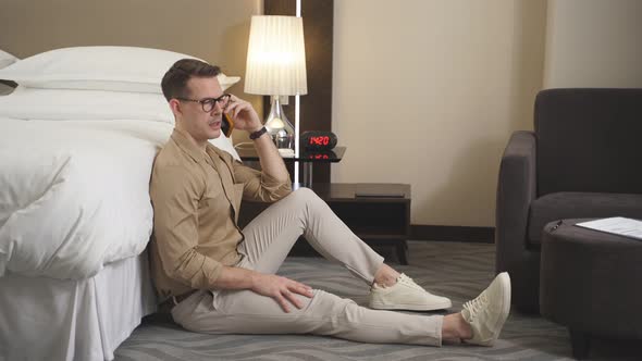 Happy Young Guy in Stylish Brown Short Sitting on Bed in Hotel and Having Talk on Phone
