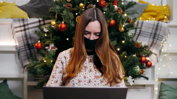 Girl in a medical mask works from home on a laptop in quarantine mode on Christmas Eve