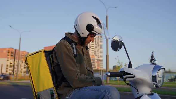A Male Courier Parked His Scooter Along the Road to See the Delivery Address on His Phone alt