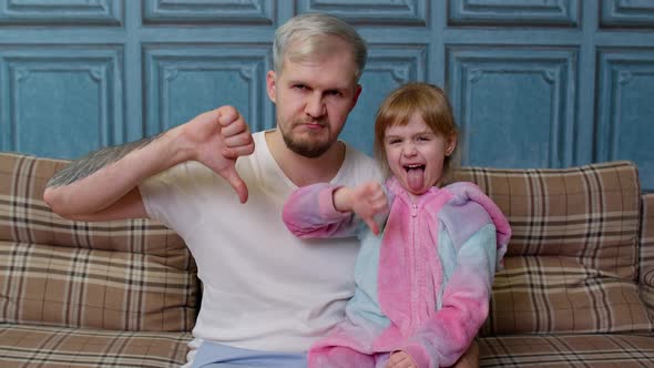 Displeased Father and Child Daughter Kid in Pajamas Sit on Couch in Room Showing Thumb Down Dislike alt