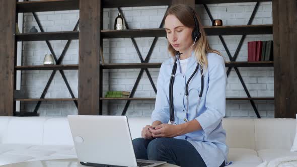Woman Wears a White Coat and a Headset While Talking on a Laptop Computer Using an Online Video Call alt