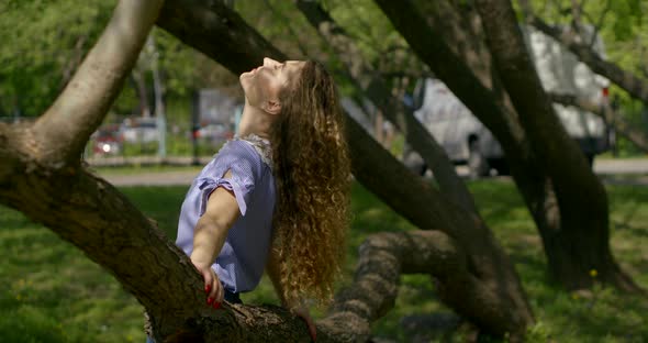 Happy Blonde Woman and Is Leaning on Tree in Park, Smiling for Camera and Touching Hair alt