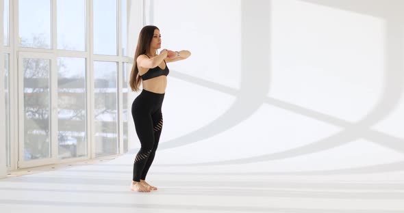 Side View of Young Caucasian Woman Working Out Doing Exercises in a White Studio with a Large Window
