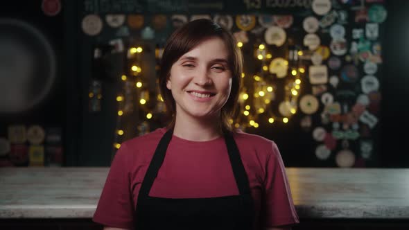 Portrait of Waitress in Apron Smiling at Camera with Hands Crossed Standing in Bar alt