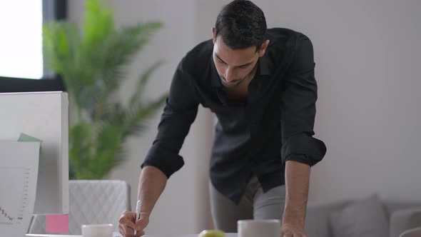 Focused Young Man Standing at Computer Table Writing Down Ideas alt