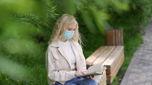 Young Beautiful Woman in a Medical Mask Reads a Book on a Park Bench
