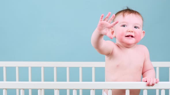 Happy infant baby boy with raised hand gesture in crib, studio blue background