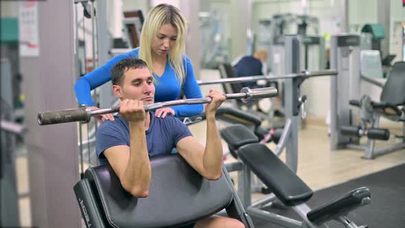 Skinny guy doing biceps exercise under the supervision of a trainer in a gym alt