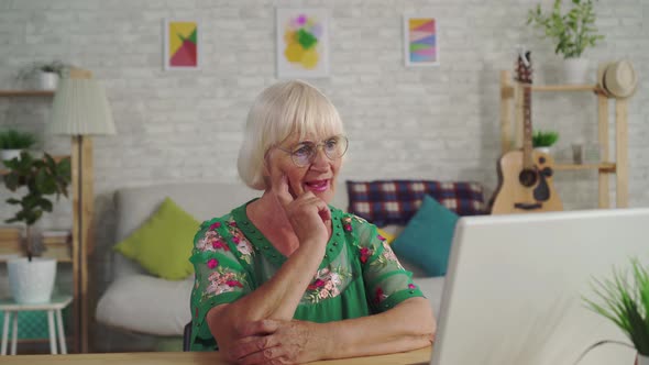 Thoughtful Older Woman Finds a Solution and Uses a Laptop Sitting at a Table in the Living Room alt