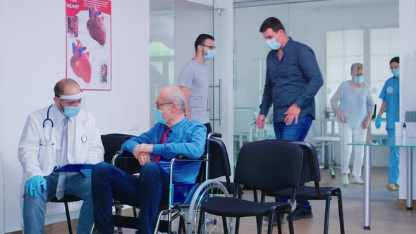 Invalid Old Man with Face Mask Discussing with Doctor in Hospital Waiting Area alt