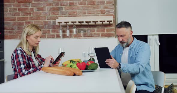 Woman Uses Phone and Sitting at Kitchen Table with Her Concentrated Bearded Husband alt