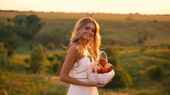 Beautiful sexy blonde girl in white dress posing in a field at sunset with a basket of fruit	 alt