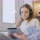 Young woman sitting by the window at home in headphones, working on a tablet. Remote study, work. - VideoHive Item for Sale