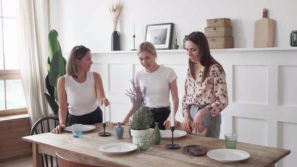 Three girls chat smile stand in the kitchen and lay out plates for a joint dinner alt
