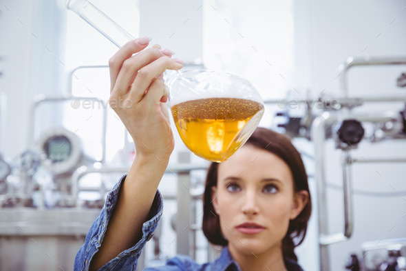 Stylish brunette in denim jacket looking at beaker of beer in the factory