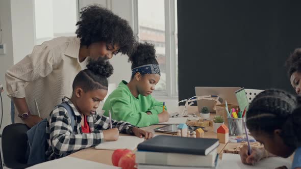 African American female teacher standing with pupils teaching writing lesson in modern classroom. alt