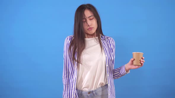 Young Sleepy Asian Woman Yawns and Stands on a Blue Background with a Up of Coffee alt
