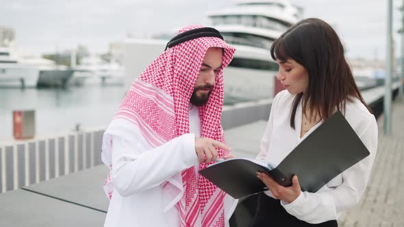 Young European Business Woman Yacht Broker Showing Contract Details to Arab Man Client in Port alt