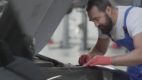 Adult Mechanic Standing Near Car with an Open Hood and Tightens the Bolts with Wrench in Engine alt