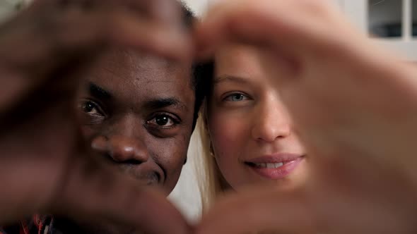 Close Up Smiling Multiethnic Couple Faces Looking Through Joined Fingers Heart alt