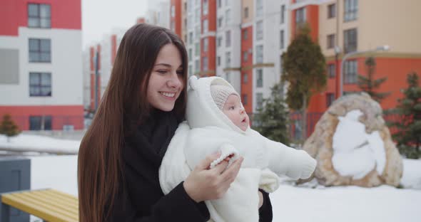 Joyful Young Mother Walks Down The Street In Winter, Holding A Baby
