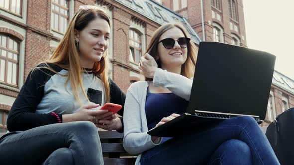 Modern Female Students Working Together on a Laptop on a Student Project While Sitting on Bench in alt