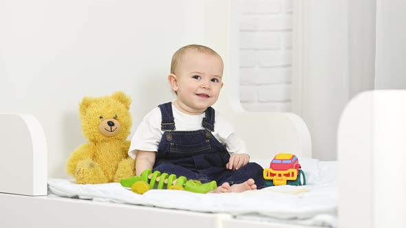 a Baby Boy in a Denim Jumpsuit Smiles and Plays with Toys While Sitting on a Bed alt