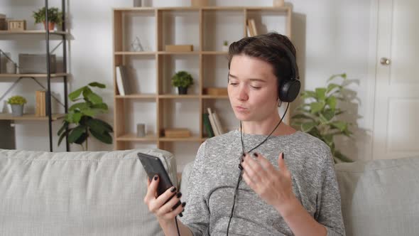 Young Woman Wear Headphones Sit on Couch at Home Communicates with Family Use Smartphone and alt