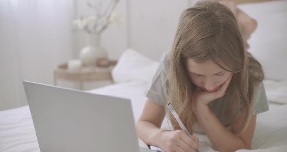 School Girl Is Lying on Bed with Laptop and Copybook, Writing Homework and Looking on Display of alt