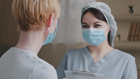 Unrecognizable Caucasian Woman Patient with Short Hair in Medical Mask Speaks to Female Doctor Nurse alt