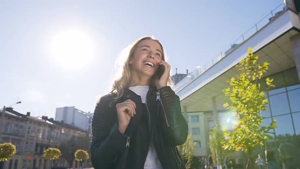 Blonde Girl Talking on Smartphone and Smiling while Walking on the Street alt