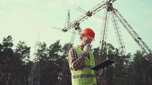 Caucasian Engineer in Protective Uniform and Orange Hard Hat Holds Walkietalkie and Clipboard on alt
