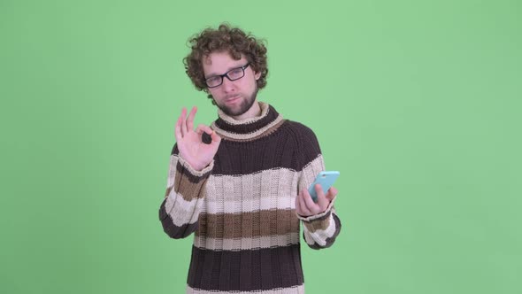 Happy Young Bearded Man Using Phone and Giving Ok Sign alt