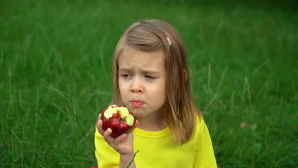 Portrait of Little Girl Sits By Green Lawn in Summer Garden and Eats Red Ripe Apple