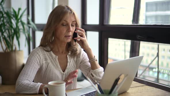 Businesswoman Having Mobile Call in Front of Laptop Screen at Table in Home Office alt