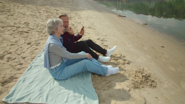 Mature Couple Sitting on Sand Listening to Music Via Smartphone alt