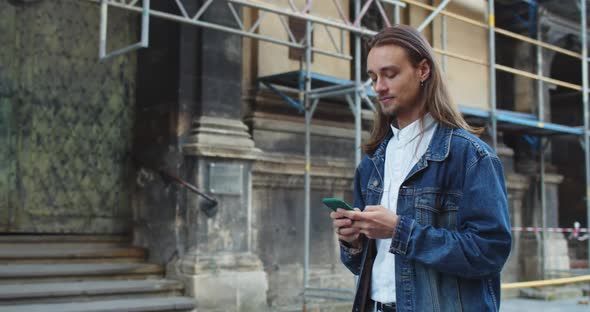 Side View of Long Haired Young Man Typing Message and Touching Smartphone Screen While Walking at alt