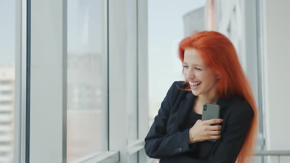 Woman Looking at Camera and Laughing. Red-haired Girl Stands By the Window in the Office and Laughs alt