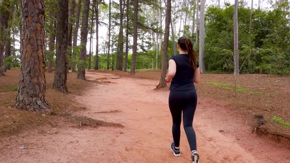 Girl jogging on a dirt trail between trees, camera following the subject from behind. alt